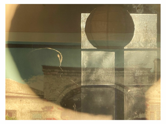 The photo shows a composition of different forms and layers. It was taken against the image of a building that is behind glass. The sun is shining inside from the back so that the window the lamp and my silhouette are visible inside in the reflection. At the same time the round form of the lamp is reproduced in the actual darker and bigger shadow of the lamp against the wall.