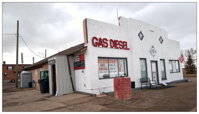 A small, white, single-story building prominently labeled "GAS DIESEL." The architecture of the building is simple with minimalistic detailing aside from a couple of diamond-shaped decorations on the facade. There's a front entrance with a brown door slightly ajar and an adjacent window displaying signage with various prices for gas and diesel, and tabs like "CASH DEBIT," "VISA MC," indicating accepted forms of payments. A red brick podium-like structure is visible by the entrance along with a couple of garbage bins and a bench beside the building.

The overall environment appears somewhat deserted or in a less maintained condition, suggesting it could be in a rural or less-urbanized location. Cloudy skies and the damp ground suggest recent or prevailing overcast, cloudy weather conditions. There are no visible people or much activity in the immediate vicinity within the frame.