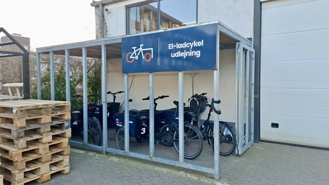 Around the back of a Silvan hardware store in the centre of Aarhus, Denmark. Paved area with wooden pallets stacked up. In a bike shed, a row of cargo bikes with the Silvan logo on them. A sign with a cargo bike logo says 'el-ladcykel udlejning'. 'Electric cargo bike rental.' On the cargo bikes themselves, it clarifies that you can borrow them for free at first, then pay for longer hours.
