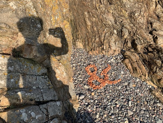 An ampersand has been made on a stony beach, using only old bits of worn away red brick, contrasting against their otherwise mostly grey brethren. The shadow of the photographer is visible on a couple of adjacent rocks.
