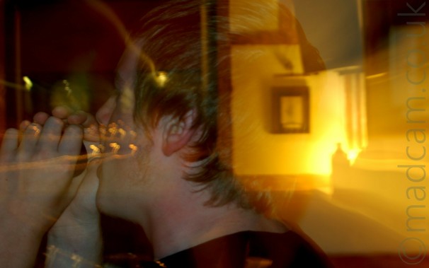 Long exposure close-up of the side of a young man's head facing to the left.
He has short blond hair, and is wearing a black t-shirt in his hands which is raised to his mouth as he sings into it.
A snap zoom has been used, starting zoomed right in, then zooming out, meaning that the background, including a picture in a frame on the wall, appears to be visible through the back of his head, lit by yellow light.
Yellow light trails are visible in the dark background on the left of the frame.