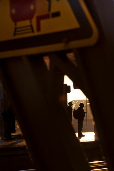 A person, visible as a mere silhouette, waiting for a train in the golden morning sun. The photo is taken through an opening in a steel beam. In the background, a bridge is visible. On the top of the frame, a warning sign.