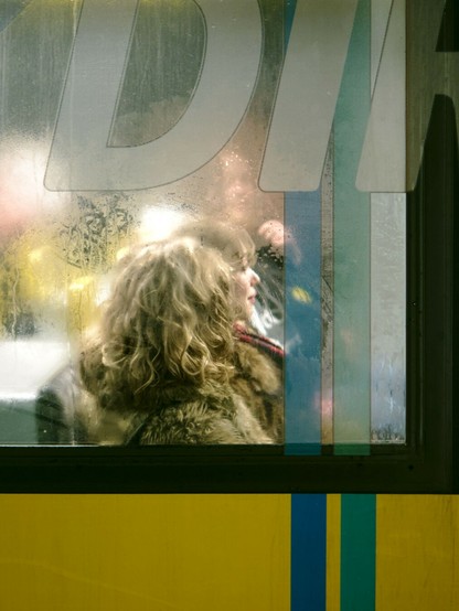 A commuter on a Berlin tram, photo taken from outside through a fogged window. The commuter's blond hair stands out. At the top, letters printed on the window are visible.
