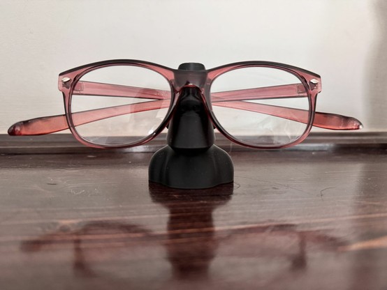 Fotografía en color de un soporte para gafas impreso en 3D con forma de moái de la Isla de Pascua. Con unas gafas de montura roja sobre su “rostro”, la figura parece observar el mundo con una expresión inesperada, fusionando el pasado ancestral con un toque moderno y lúdico. La escena transcurre sobre una mesa de madera oscura, con reflejos sutiles y un fondo neutro.

Color photograph of a 3D-printed glasses holder in the shape of an Easter Island moai. With a pair of red-framed glasses resting on its “face,” the figure appears to gaze at the world with an unexpected expression, blending ancient heritage with a modern, playful touch. The scene is set on a dark wooden table, with subtle reflections and a neutral background.