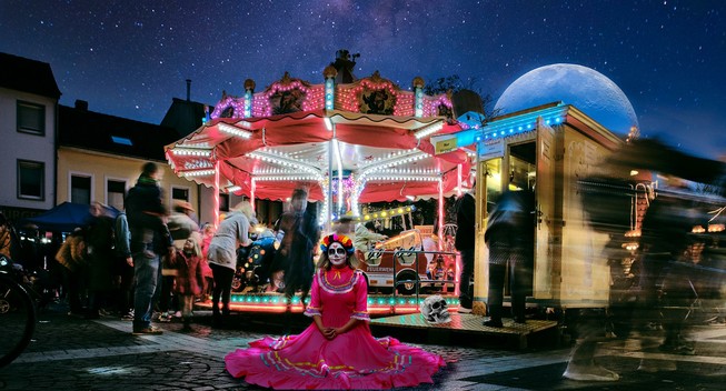 A small illuminated carousel at night on a market square. A starry sky above with a huge moon rising above the carousel. In the foreground a woman in a flowing pink dress with Mexican Day of the Dead makeup sits in front of the carousel with the dress spread out around her on the ground.
