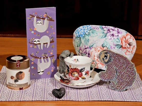 Peach and lavender tea tableau with a sloth theme featuring adorable sloths on a greeting card, as a figural teapot infuser, and a sparkly soft sculpture that is actually a dish scrubber but too cute to use. There is vintage china, a blown glass heart ornament, and a floral handmade cozy cover, and the tea is Tangerine PuErh black tea from the Tao of Tea.
