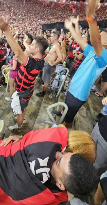 Fotos de pessoas em uma arquibancada. A pessoa que segura o celular esta tomando uma self dele com uma mulher sentada sobre ele com a bandeira do Flamengo tapando-os.