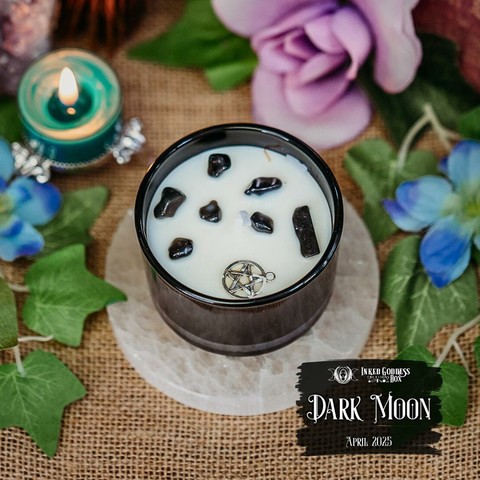 A white candle in a black tin sits on a selenite disc. The candle has several dark gemstones and a silver pentacle charm embedded in the surface. This is one of the items in the exclusive April 2025 "Dark Moon" Inked Goddess Creations Box.