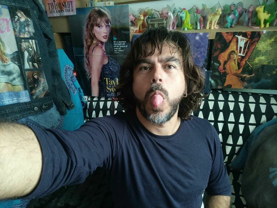 Selfie pulling my tongue out. Background is the studio where I record my videos for YouTube, lots of pictures of Taylor Swift.