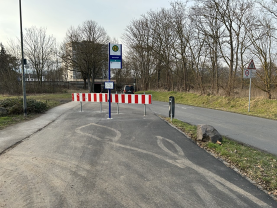 Straße mit linksseitigem Hochbord. Direkt vor der Kamera breite Barrieren über die gesamte Fahrbahnbreite, weiterhin in der mitte ein Mast mit Bushaltestellenschild und Fahrplänen. Rechts ein Abhang mit einem kleinen Schildermast und einem Fels. Links neben dem Hochbord eine Straßenlampe und ein Gebüsch. Auf der Straße sind Reifenspuren eines PKW, der offenbar gewendet hat.