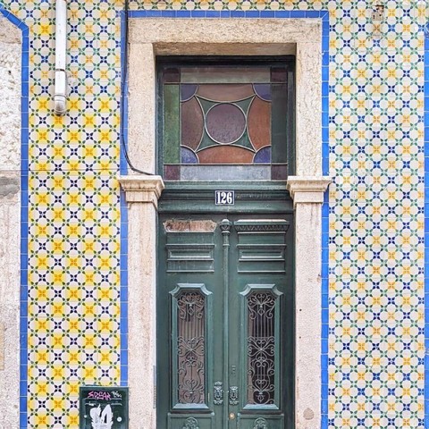 A green door with decorative metalwork and a square stained glass window above, set in a tiled building with the number 126.