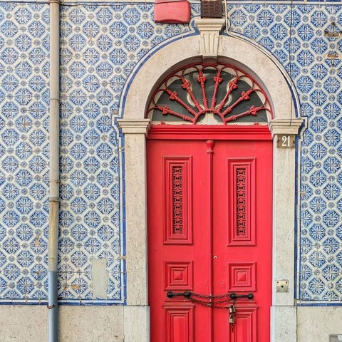 A bright pink double door under a stone arch with a decorative fanlight, set in a building with blue and white patterned tiles. The number 21 is visible to the right. The door is locked with a padlock and chain.