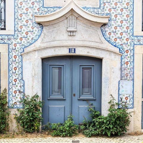 A pale blue double door with small windows, under a stone arch with the number 18, set in a tiled building with weeds growing in front of the door.