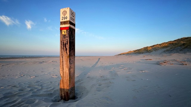 A wooden beach pole is gently illuminated with golden light at sunset. A sandy beach, the North Sea and dunes with marram grass in the background.