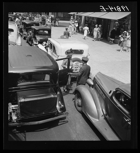 The image depicts a bustling street scene in an urban environment. Multiple vintage cars are visible, with one prominently displayed on the right side of the frame and another partially obscured behind it to the left. There is a clear view of the car's shiny exterior and distinctive design features such as rounded fenders, large chrome bumpers, and wire-spoke wheels.

On the street, pedestrians dressed in early 20th-century fashion are walking or standing near storefronts with awnings extended over the sidewalk. The clothing styles suggest women wearing dresses and hats typical of that era, while men appear to be wearing suits and fedoras as headwear.

A person is seated on a motorcycle parked amongst the cars, adding another layer of activity to the scene. This individual appears engaged in an interaction or conversation with someone standing next to them, possibly inside one of the vehicles.

The overall atmosphere captures a moment from daily life in what could be New York City during the 1930s-40s era, as indicated by the clothing styles and vintage automobiles present. The image conveys a sense of movement, social interaction, and economic activity characteristic of that time period's urban environment.

The photograph is black-and-white, which enhances its historical context but also limits color information for analysis or artistic purposes beyond those related to form, texture, and composition.