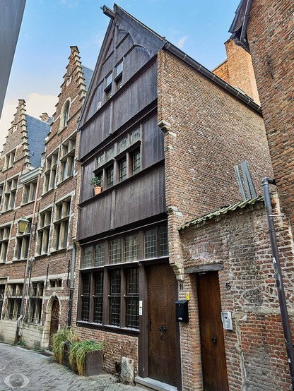 A low-angle shot looking up at a narrow street lined with old brick buildings. On the right is a tall, narrow building with a wooden facade on the second and third stories. There are rows of windows with many panes of glass on each floor. A small plant sits in a pot on the second floor. Next to the building is a brick wall with a small door and the number "15" on it. To the left of the wooden building is another brick building with a more ornate facade and many windows. The sky is blue with some clouds visible in the upper left corner of the image.