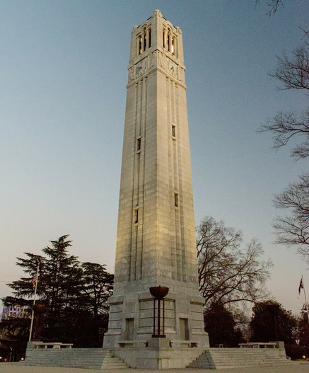 A photo of the NCSU belltower on Hillsborough street during sunset, partially illuminated with a darkening gray-blue sky behind it.