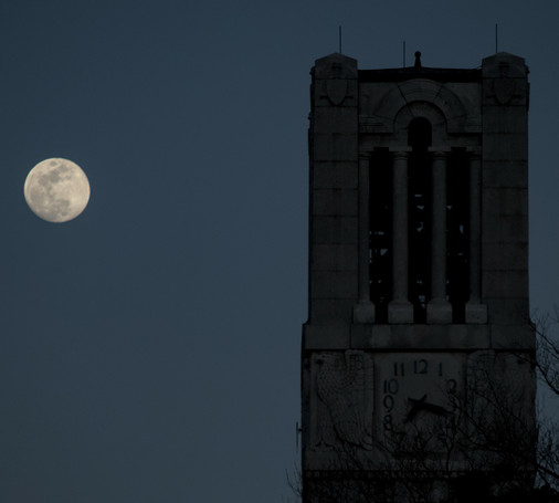 A photo of the top of the belltower with an almost full moon behind it.
