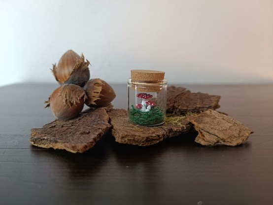 A tiny glass with a miniture of a fly agaric mushroom inside is standing on a piece of bark.
Besides it lie three hazelnuts. The glass is about the size of 1 1/2 nuts.