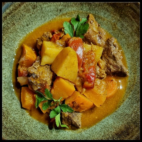 Stücke von Kürbis, gebratenem Rind, Paprika, Kartoffeln,  Möhren in einer orangenen Sauce, ein paar Korianderblättchen; auf einem grünlich glasierten Keramikteller