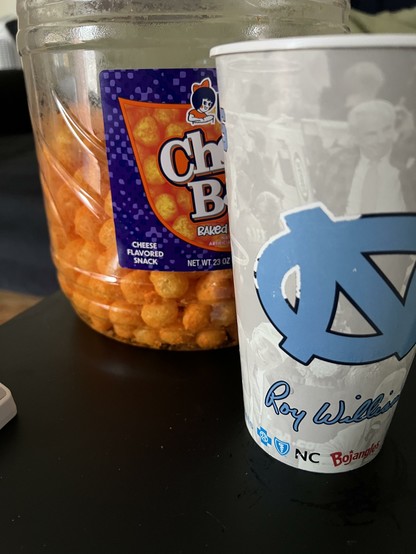 A tall, well-worn North Carolina Basketball souvenir cup with Roy Williams theme next to a giant half-empty canister of Utz cheese balls for March Madness game watching