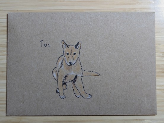 A recycled paper envelope with the word "To", followed by a pen & coloured pencil sketch of a dingo puppy sitting down with its legs sprawled under it