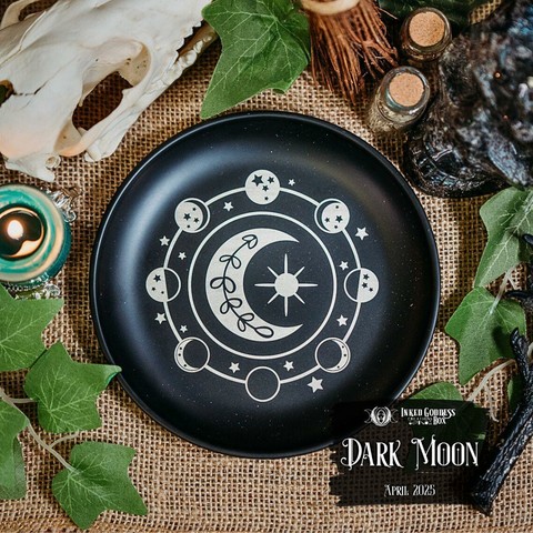 A small black plate sits on a burlap-covered surface. The plate has a light-colored circle of moon phases and stars painted on it, with a crescent moon and small sun in the center. This is one of the items in the exclusive April 2025 "Dark Moon" Inked Goddess Creations Box.