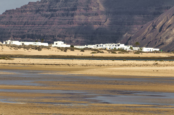 una grande spiaggia con bassa marea e alcune piccole casette. Sullo sfondo, scogliere altissime in lontananza.