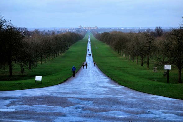 A distant shot of a long, straight path leading to a castle in the distance. The path, wet and reflective, stretches through a park-like setting with bare trees lining both sides. People are walking along the path. The sky is overcast and grey. The perspective suggests the photo was taken from a slightly elevated position.