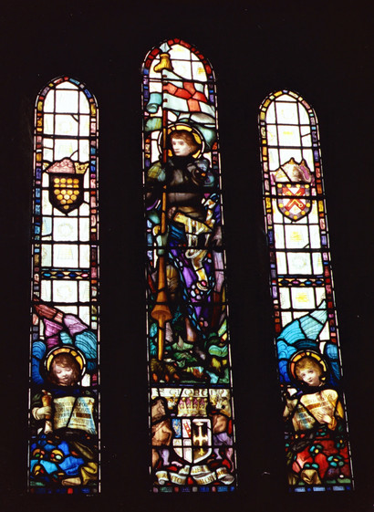 Stained glass window from St. Materiana's Church, Tintagel. It has three panels, with the left panel depicting Cornwall Coat of Arms (15 gold coins) with an angel reading a scroll below. The right hand panel has an unknown ( to me) crest with a crossed key and sword, with an angel below reading a scroll. The centre panel features St. George in armour holding a lance with his Pennon flying (red cross on white background). He is stood over a dragon, and the bottom of the panel contains another crest flanked by a horse and a deer (possibly).