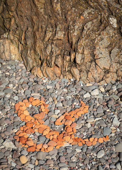 An ampersand created by arranging old bits of red brick, worn and rounded by the sea, against other less colourful stones on a rocky beach.