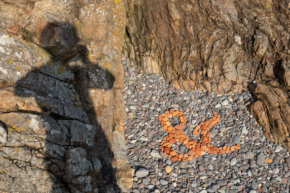 An ampersand created by arranging old bits of red brick, worn and rounded by the sea, against other less colourful stones on a rocky beach. The creator's shadow is visible on the adjacent rocks, as he photographs his work.