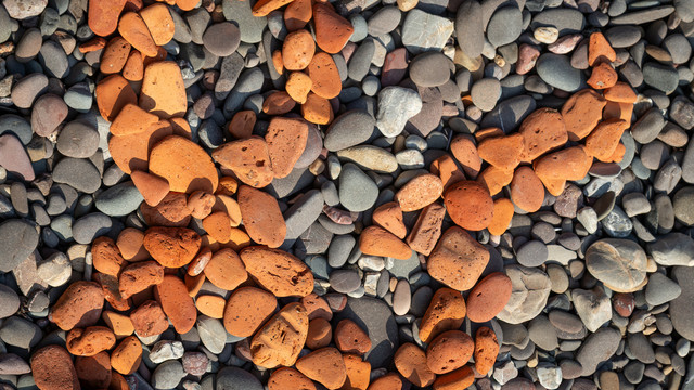 A close-up detail of part of an ampersand created by arranging old bits of red brick, worn and rounded by the sea, against other less colourful stones on a rocky beach.