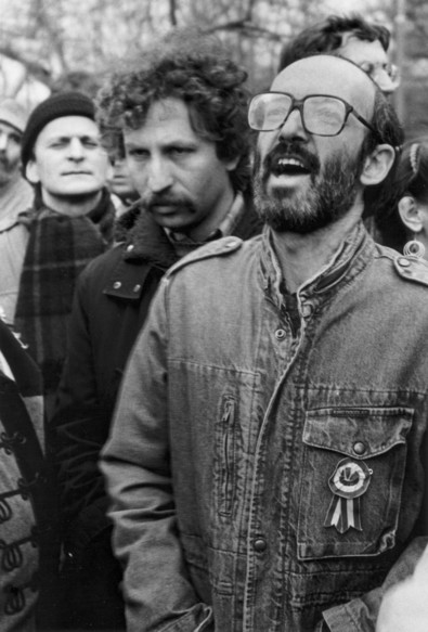 A monochrome photo of a man wearing a denim-like jacket surrounded by people.  On his left breast is a cockade than would have been in the Hungarian national colours of red, white, and green. His head is titled slightly backwards and his mouth is open suggesting he is speaking loudly.