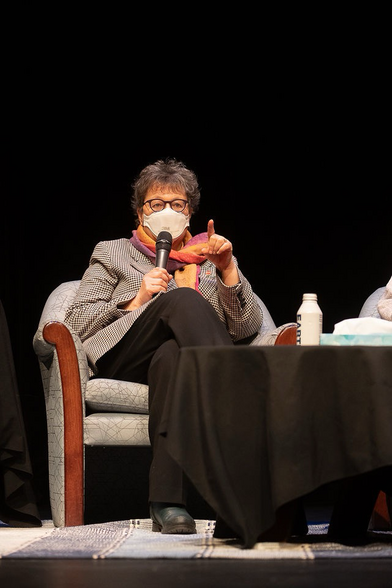 Senator Simons, in a black and white houndstooth blazer that her husband gave her for Christmas , black slacks, rainbow scarf and white N95 face mask, sits on the stage of the Timms Theatre, a microphone in