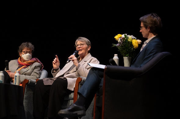 Julie Lloyd, centre, in a natural silk blazer, tells a story, holding a microphone,, while Senator  Simons and Dean Fiona Kelly listen attentively.