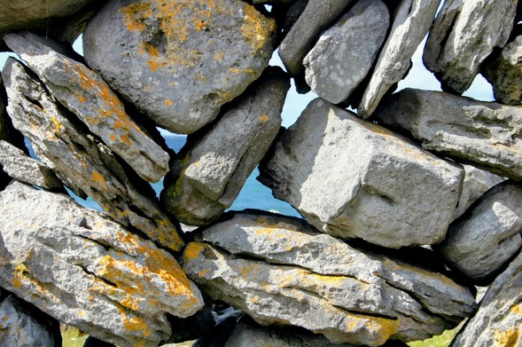 Eine Mauer, locker zusammengesetzt aus unterschiedlichen großen und geformten hellen Steinen mit gelben Flechten. Durch die Steine hindurch schimmert blau das Meer. In der Mitte des Bildes sieht man einen dreieckigen Durchguck.