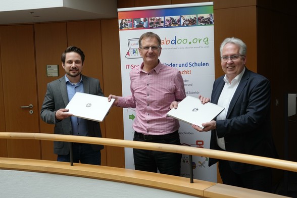 In einem Bürogebäude stehen 3 Männer auf einem Flur, im Vordergrund eine Brüstung mit heller Holzreling, im Hintergrund eine etwas dunklere Holzvertäfelung. 
Links steht Patrick Wiedemeyer, Stabsbereichsleiter IT und Digitalisierung, in der Mitte Armin Cibulski von Labdoo und rechts Landrat Jürgen Müller vom Kreis Herford. Im Hintergrund steht ein Roll-Up von Labdoo. Je 2 Personen halten eine silbernen HP-Laptop zwischen sich. Alle schauen in die Kamera und lächeln.