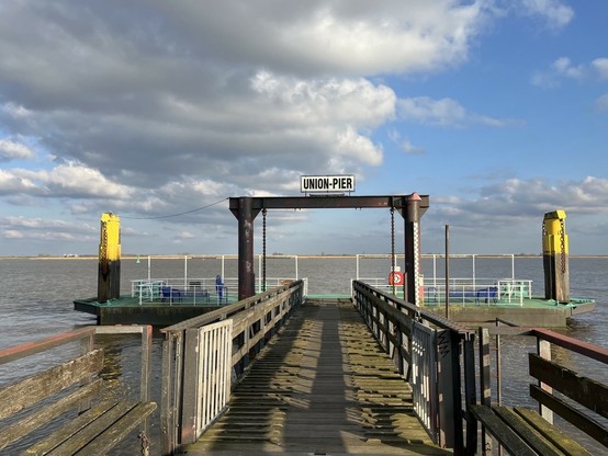 Das Bild zeigt einen Anleger, bezeichnet als „UNION-PIER“, an einem ruhigen Gewässer. Ein Steg führt von der Kameraposition aus zum Anleger, der mit einer einfachen, aber robusten Konstruktion aus Metall und Holz ausgestattet ist. Rechts und links befinden sich gelbe Bojen. Auf dem Anleger befinden sich einige einfache Sitzgelegenheiten. Der Himmel ist leicht bewölkt, aber es scheint sonnig zu sein.  Das Bild vermittelt einen Eindruck von Ruhe und Abgeschiedenheit.
The picture shows a jetty, labeled "UNION-PIER", on a calm body of water. A jetty leads from the camera position to the pier, which is equipped with a simple but robust construction made of metal and wood. There are yellow buoys to the right and left. There are a few simple seats on the jetty. The sky is slightly cloudy, but it appears to be sunny.  The picture conveys an impression of peace and seclusion.