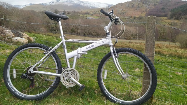 A scruffy Dahon Expresso folding bike leaning on a wire fence in front of Welsh hillside.