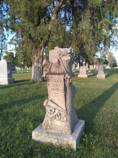 A cemetery with green grass and trees bathed in afternoon sunlight. The focus is a gravestone which is carved to look like a tree stump. There are a few stumps of limbs hewn close to the trunk, and a smooth section for the inscription appears to be hung by a carved rope. M. Peck was born in 1801 and died in 1890.