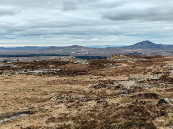 A boggy and boulder strewn landscape stretches away to the sea, framed between two distant mountain humps. The sky is mostly covered with grey clouds.