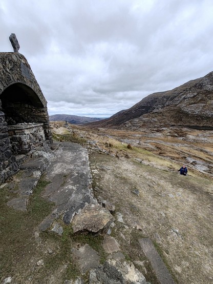 Picture of a stone altar in a small arched stone shelter, with a view over a mountain pass, and down a valley into the distance. A rocky mountain slope rises from the right hand side. The sky is overcast.