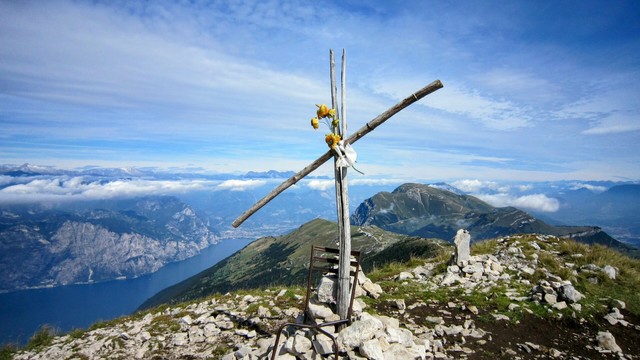 A wide shot from a slightly low angle shows a wooden summit cross atop a rocky mountain peak. The cross is made of roughly hewn wooden beams, with a vertical post and a shorter horizontal beam. It's adorned with a few yellow flowers and a white cloth tied to it. The cross is mounted on a metal frame that is secured amongst the loose grey rocks of the summit.
Behind the cross, a stunning vista unfolds. Rolling, green and rocky mountain ranges stretch out towards the horizon, some with visible hiking trails winding along their slopes. In the distance, a large, deep blue lake snakes through the valleys, partially obscured by low-lying clouds that fill some of the lower areas. The sky above is a mix of clear blue and scattered white clouds. The overall impression is one of a high-altitude, scenic viewpoint on a bright day.