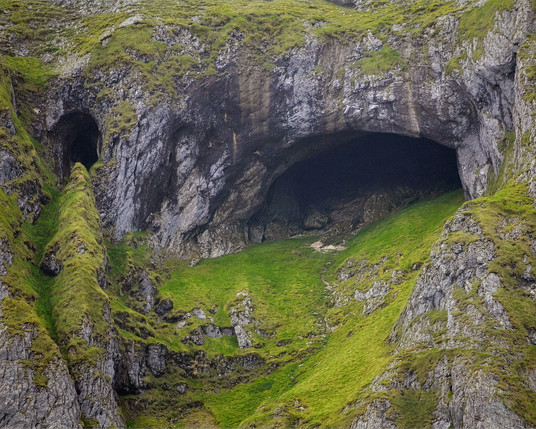 Diarmuid and Grainne’s Cave, nestled high in the rugged terrain of Benwiskin mountain in County Sligo, Ireland. The cave features two prominent entrances, framed by dark, textured rock that contrasts sharply with the surrounding lush green grass. The slope leading to the cave is dotted with patches of grass and rocky outcrops, creating a dramatic landscape. The overall scene evokes a sense of ancient mystery and natural splendour, with the cave openings suggesting depth and intrigue against the mountainous backdrop.
