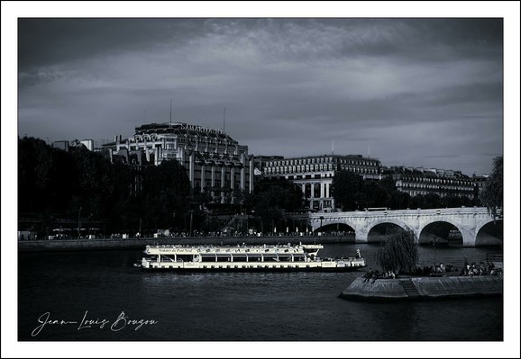 In this evocative image, a tranquil scene unfolds along the banks of a river, the Seine, given the context. The black and white palette adds a timeless quality, accentuating the stark contrasts between light and shadow.
A gently cruising boat, illuminated by warm lights, stands out against the cooler tones of the landscape, suggesting a sense of life and movement amid the stillness. The reflections on the water create a shimmering texture that draws the eye, inviting one to linger on the scene.
In the background, stately buildings rise gracefully, their architectural details softened by the monochromatic scheme. The presence of trees adds a touch of nature, balancing the man-made structures and hinting at a connection between urban life and the natural world.