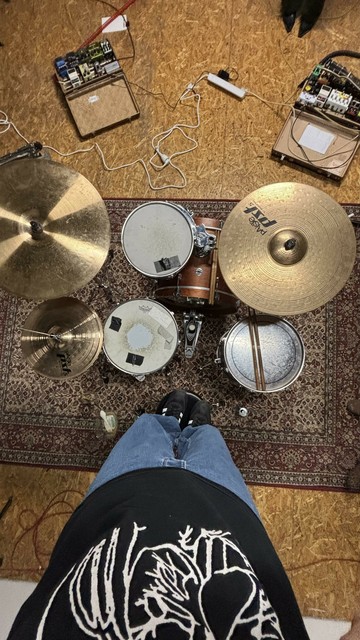 An overhead view of a drum set with two cymbals, a floor tom, a snare drum, and a bass drum. Various pedals and a tangle of cables are visible on the ground, along with a decorative rug