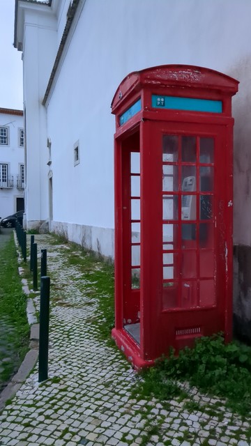 Cabine telefónica vermelha, num passeio de calçada, encostada a uma parede branca.
Vêem-se muitas ervas daninhas por todo o passeio.