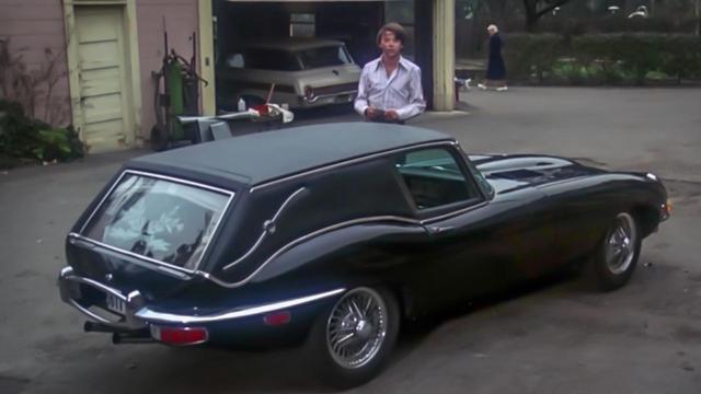 A still from the 1969 film Harold and Maude. Harold is looking towards the camera with a garage behind him. In front of him is his black e-type Jaguar which he modified to function as a hearse.