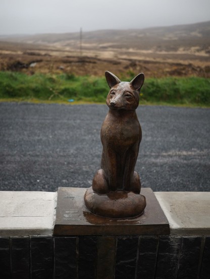 A bronze statue of a fox with a misty, early spring landscape in the background.